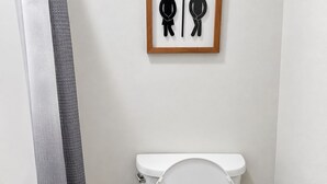 Une salle de bain moderne avec des toilettes blanches, un panneau indiquant les toilettes pour tous, une plante en pot et un tapis texturé gris.
