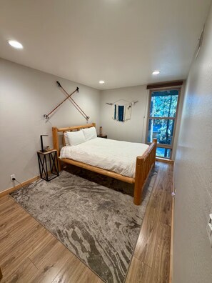 Ein Schlafzimmer mit einem hölzernen Bett, einem kleinen Nachttisch, einer Wandleuchte und einem Fenster mit Blick auf Bäume.