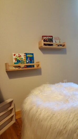 Quarto com um banquinho branco e fofo, estantes de madeira com livros infantis e parede branca.