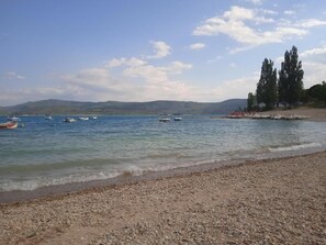 A pebbled beach with calm water, boats, and distant hills.