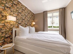 A hotel room with a floral wallpaper, a bed with white bedding, a bedside table with a book, and a window with curtains. - 5, Residence Dunenstrasse (Binz)