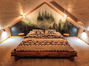 Un dormitorio con techo inclinado, una cama con ropa de cama estampada, dos lámparas fijadas a la pared y un mural con motivos de bosque.