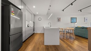Une cuisine moderne avec un réfrigérateur en acier inoxydable, un îlot central blanc doté d’un évier, un plancher en bois et un coin repas avec un canapé vert et une table.