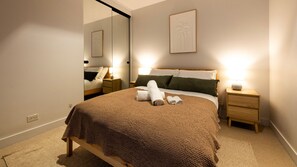 Quarto de hotel com uma cama, duas mesinhas de cabeceira, um criado-mudo, um espelho grande e duas obras de arte emolduradas na parede.