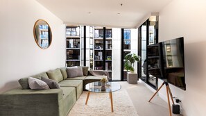 A modern living room with a sectional sofa, a glass coffee table, a large flat-screen TV, and a bookshelf. - Eighteen Above Docklands Skyline & Pool (Docklands)