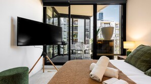 A hotel room with a large flat-screen TV, a bed with a green pillow, a bedside table with a lamp, and a view of the city through a large window. - Eighteen Above Docklands Skyline & Pool (Docklands)