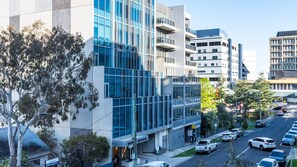 A multi-story building with a glass facade, a parking lot with several cars, and a street with pedestrians. - Eighteen Above Docklands Skyline & Pool (Docklands)