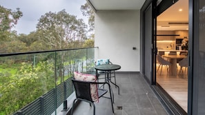 Ein Balkon mit Glasgeländer, einem Tisch mit Stühlen und Blick auf Grünflächen und Bäume.