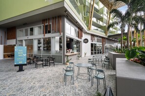 Ein moderner Essbereich im Freien mit Tischen und Stühlen, ein Restaurantschild und ein Mehrstöckiges Gebäude mit grüner Fassade.