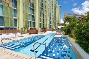 En swimmingpool med ett kaklat poolområde, omgiven av en byggnad med många fönster och en grön fasad.