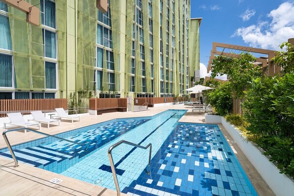 Ein Schwimmbecken mit Fliesenrand, umgeben von einem Gebäude mit vielen Fenstern und einer grünen Fassade.