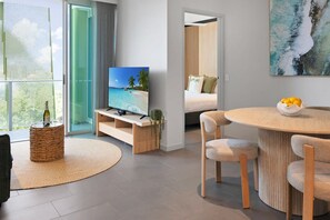Ett modernt hotellrum med en tv, en matplats med ett runt bord och stolar, en säng med utsikt över stranden och en balkong med en stol av rotting och en flaska.