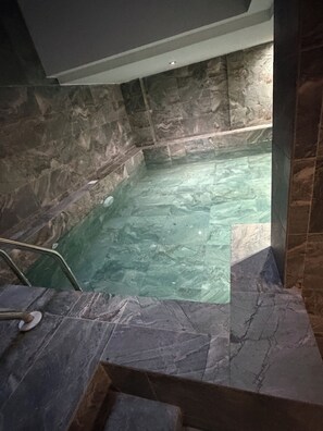 Tinaja de piedra con agua cristalina, una escalera metálica y suelo de piedra.