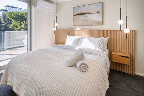 Une chambre d’hôtel avec un grand lit, une tête de lit en bois, un tableau au mur et un balcon offrant une vue sur les arbres et les bâtiments.