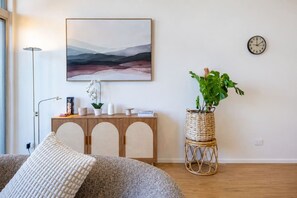 Un salon comprenant un canapé, une console ornée d’objets décoratifs, une horloge murale et un grand tableau représentant un paysage.