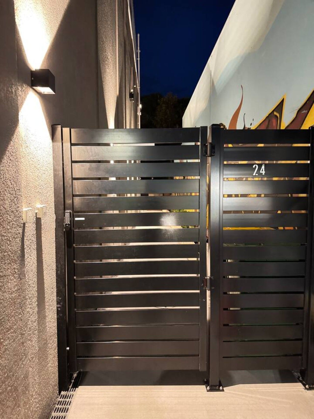 A black metal gate with horizontal slats and the number 24 on it, positioned at the entrance of a building.