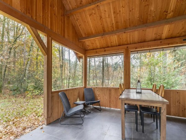 A wooden cabin with a dining table and chairs, large windows, and a view of a forest. - Beautiful bungalow with a canopy in nature (Stramproy)