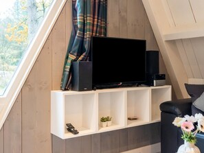 Ein modernes Wohnzimmer mit einem Fernseher auf einem weißen Regal, einer Fernbedienung, einer kleinen Pflanze und einem schwarzen Sofa.