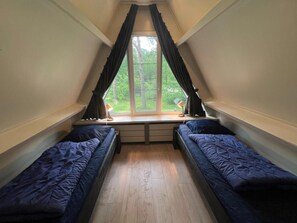 Ein Schlafsaalzimmer mit zwei Einzelbetten, einem Fenster mit Vorhängen, einem Schreibtisch mit Lampe und Blick ins Grüne.