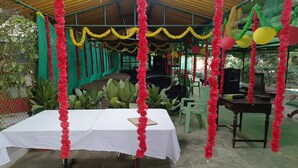 A covered outdoor area with a table set for a meal, surrounded by chairs and greenery.