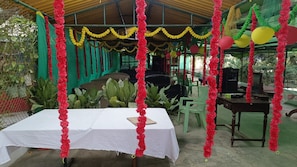 A covered outdoor area with a table set for a meal, surrounded by chairs and greenery.