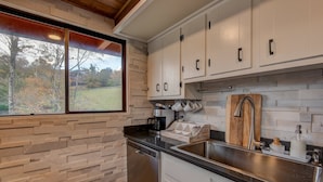 A kitchen with a stone backsplash, stainless steel appliances, and a window with a view of trees. - LUXE | SKI-IN/OUT | Hot Tub | Fire Pit | A Frame (Beech Mountain)