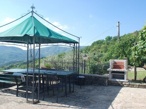 An outdoor dining area with a table and chairs under a green canopy, a barbecue grill, and a view of a mountainous landscape. - Villa in Poppi With Swimming Pool (Poppi)