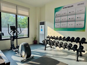 Uma academia com equipamentos de exercício, um cartaz fixado na parede com as regras da academia e uma janela com persianas.