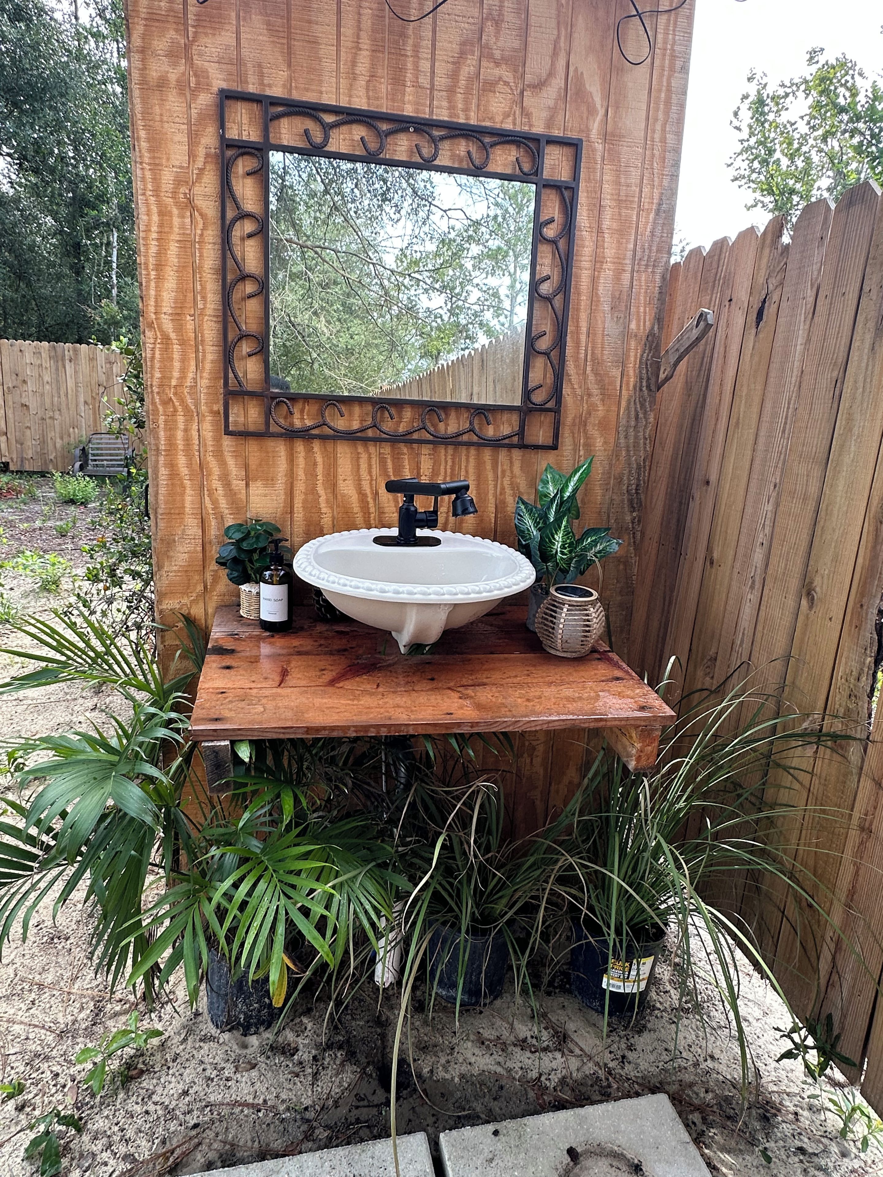 A wooden outdoor bathroom with a white sink, a black faucet, and a decorative mirror.