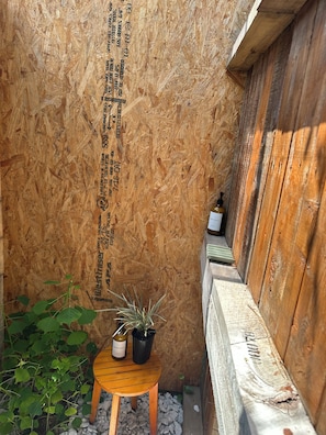 Ein schmaler Holzkorridor mit einer Holzbank, einem kleinen Tisch mit einer Flasche und einer Topfpflanze sowie einer Wand mit vertikalem Text.