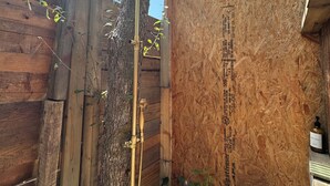 Un espace de douche extérieur en bois, avec un arbre, des murs en bois et une pomme de douche en métal.