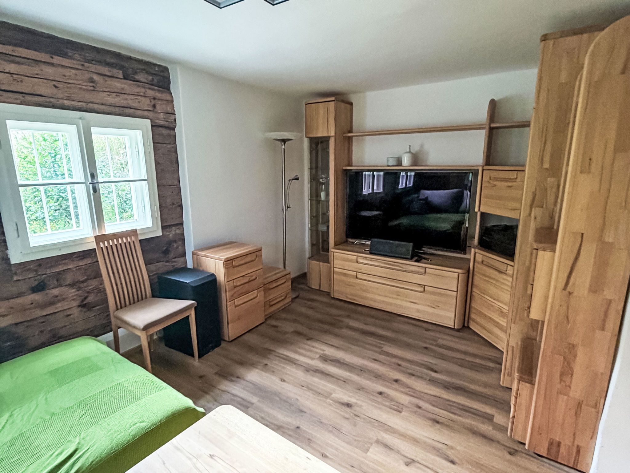 Ein kleines Zimmer mit einem Bett, einem Sessel, einem Fernseher und Holzschränken.