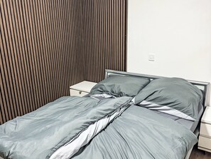 Una cama con ropa de cama gris, una mesita de noche blanca y un cabecero de madera con listones.
