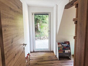 Pasillo interior de madera que conduce a una puerta de cristal con un muro de piedra al exterior. Se ve un estante pequeño con libros.