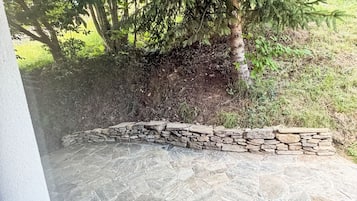 Ein gepflasterter Weg führt zu einer Steinmauer, umgeben von Grün und einem Baum.