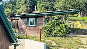 A rustic wooden house with a green window shutter, a stone wall, and a small garden area with a yellow slide.
