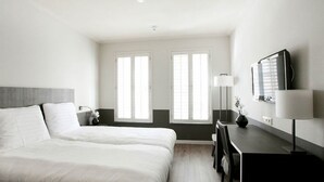 Et hotelværelse med en seng, et skrivebord, en lampe og to vinduer.