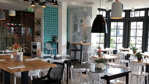 En restaurantindretning med flere borde og stole, hvoraf nogle står med planter. Der er hængende lamper og et stort vindue med udsigt.