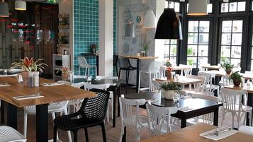 A restaurant interior with multiple tables and chairs, some with plants. There are hanging lights and a large window with a view.
