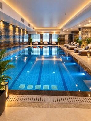 Una amplia piscina interior con agua cristalina y azul, rodeada de zonas de descanso con asientos y plantas en macetas.