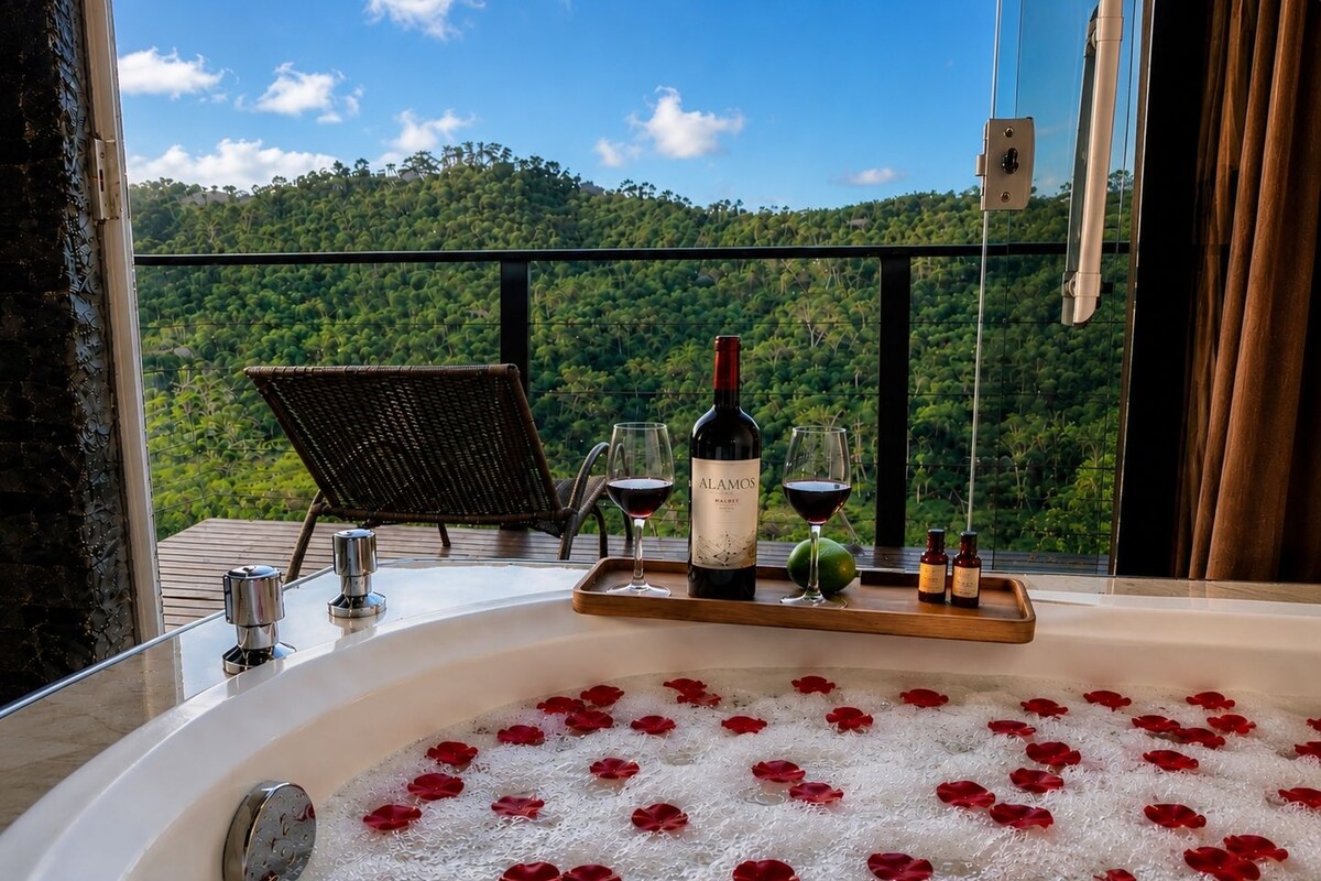 A hot tub with rose petals, a bottle of wine, and glasses on a balcony with a view of a forested hill.