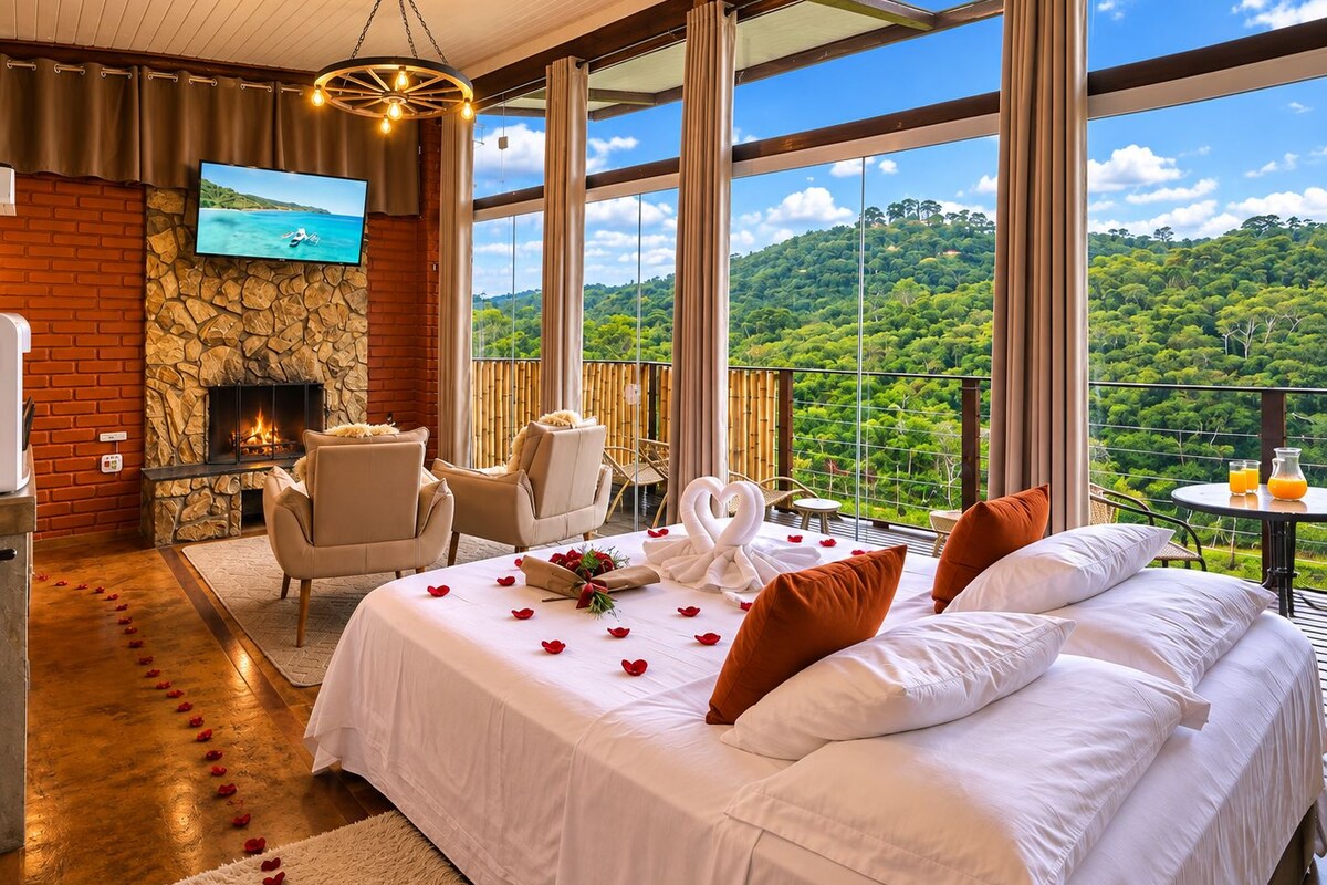 A bedroom with a bed, two chairs, a fireplace, a TV, and a balcony with a scenic view.