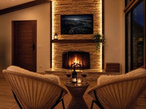 A cozy living room with a fireplace, two chairs, and a table with a bottle and glasses. - Recanto conexões  (Santo Antônio do Pinhal)