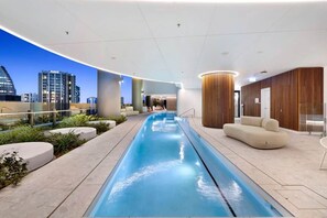 A modern indoor pool area with a long, narrow pool, seating areas, and a view of a cityscape. - Tower 2 – Broadbeach 151sqm Ocean Sky Villa 3BR (Broadbeach)