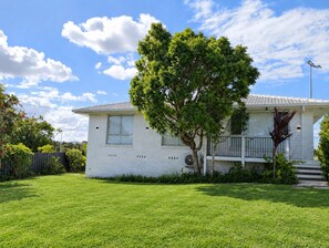 A single-story house with a white exterior, a visible air conditioning unit, a tree in front, and a clear blue sky with clouds. - Sawtell Garden House Dog Friendly Huge Yard Sleeps 6 (Sawtell)