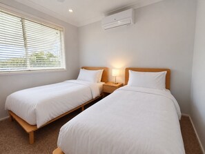 A hotel room with two single beds, a bedside table with a lamp, an air conditioning unit, and a window with blinds. - Sawtell Garden House Dog Friendly Huge Yard Sleeps 6 (Sawtell)