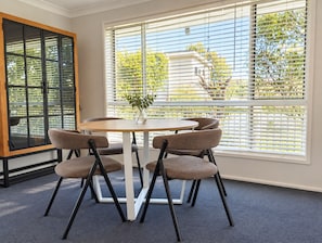 A room with a round table, four chairs, a vase with a plant, and large windows with blinds. - Sawtell Garden House Dog Friendly Huge Yard Sleeps 6 (Sawtell)