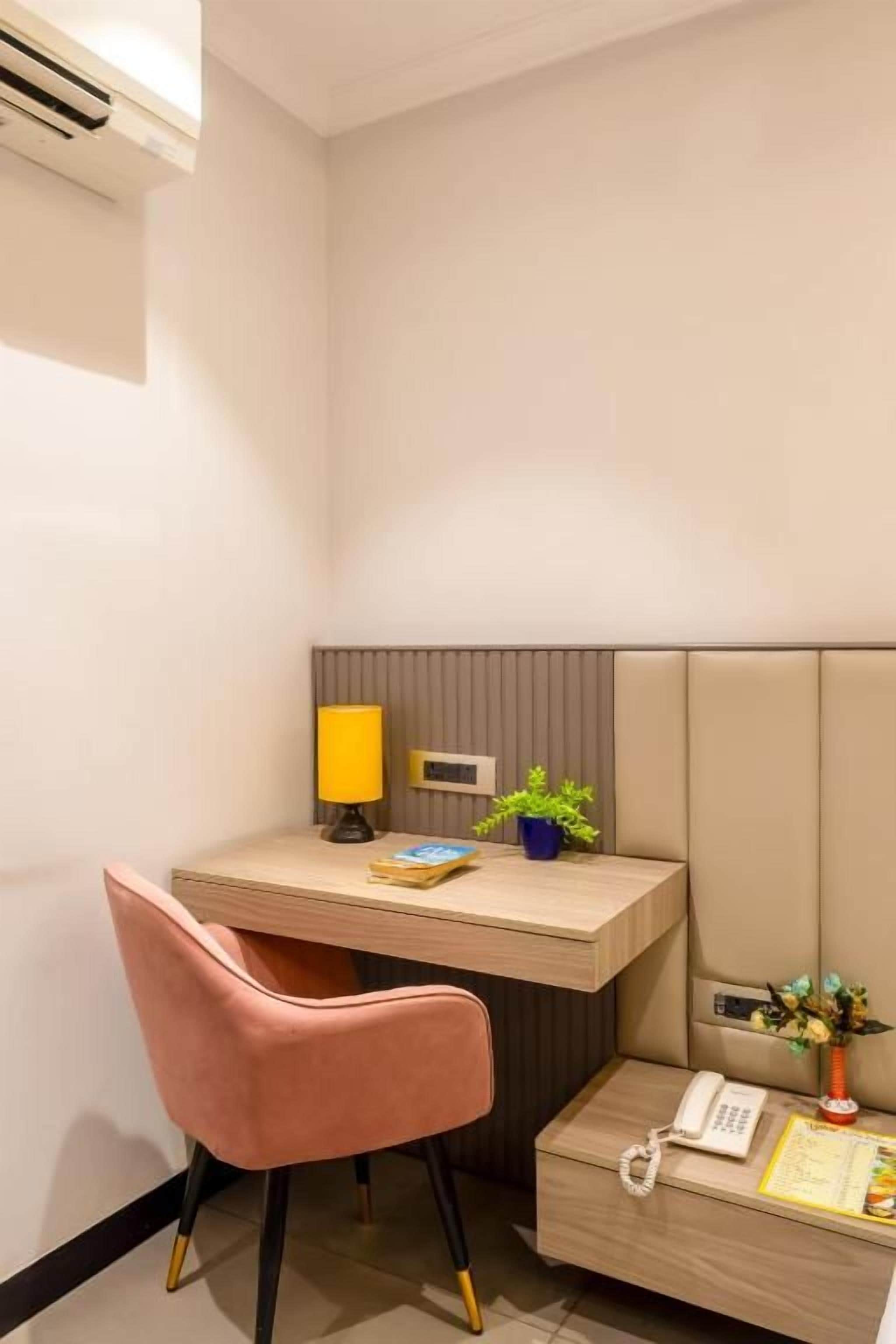 A compact hotel room with a desk, a chair, a bedside table with a lamp, a phone, and a small plant.