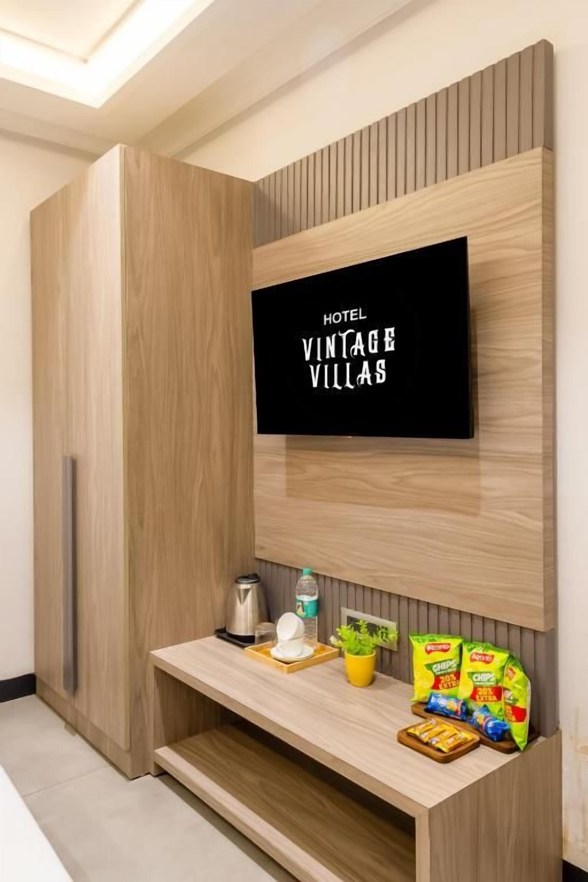 Una habitación de hotel moderna con un televisor de pantalla plana montado en una pared de madera, un pequeño estante con aperitivos, una tetera y una planta.