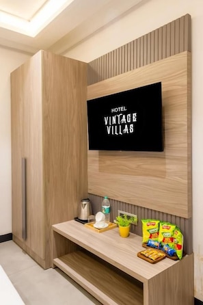 A modern hotel room with a flat-screen TV mounted on a wooden wall, a small wooden shelf with snacks, a kettle, and a plant. - Hotel Vintage Villas  (New Delhi)